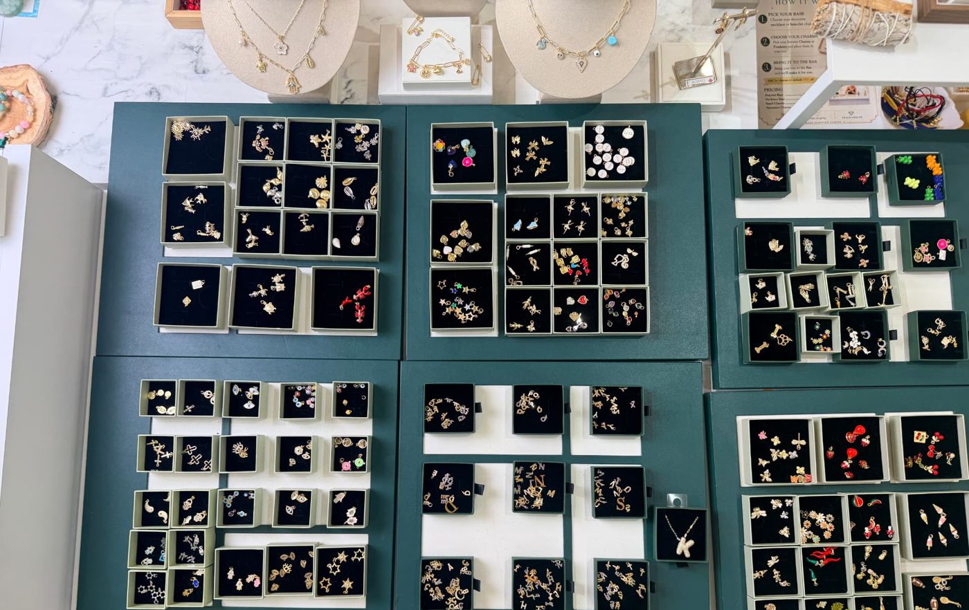 The ROMALI Charm Bar table: green trays of gold charms in small black boxes, with necklace displays behind.
